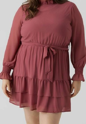 Frau trägt ein langärmliges Kleid in staubigem Rosaton mit gerafftem Saum, tailliert mit einem gebundenen Stoffgürtel und elastischen Bündchen.
