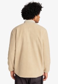 Giacca in pile beige con una superficie morbida e testurizzata, dotata di colletto e maniche lunghe. La vista posteriore mostra una vestibilità rilassata e ampia.
