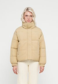 Μπεζ καπιτονέ μπουφάν με ψηλό γιακά, σχεδιασμό με ραφές και ορατά ραφές. Διαθέτει δύο πλευρικές τσέπες και ελαστικά μανίκια.