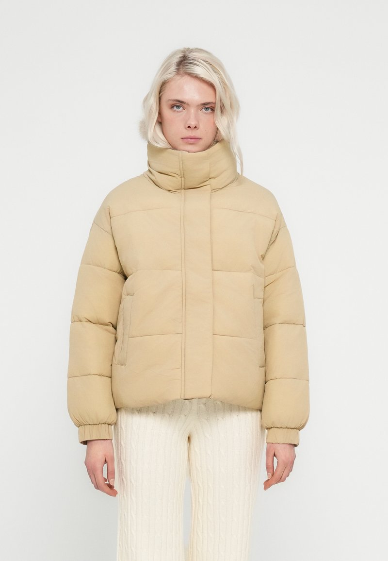 Μπεζ καπιτονέ μπουφάν με ψηλό γιακά, σχεδιασμό με ραφές και ορατά ραφές. Διαθέτει δύο πλευρικές τσέπες και ελαστικά μανίκια.