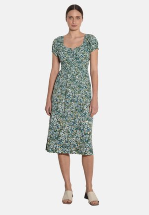 Kobieta w zielonej, kwiecistej sukience midi z krótkimi bufkami i kwadratowym dekoltem, w połączeniu z beżowymi sandałami na słupku z odkrytymi palcami.