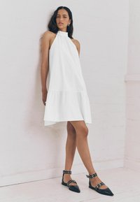 Abito bianco a collo halter con orlo a strati, presenta una vestibilità comoda e una texture liscia. Scarpe basse nere con punta appuntita, cinturino e dettagli in metallo.