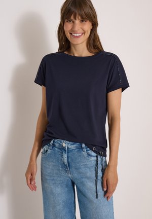 Femme souriante portant un haut à manches courtes bleu marine avec garniture aux épaules et un jean bleu clair avec un détail à cordon sur le côté droit.
