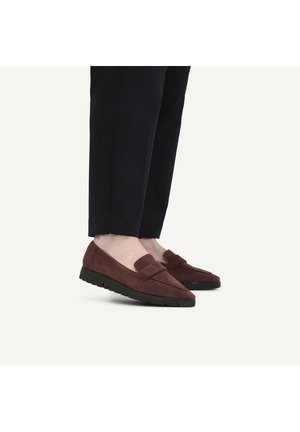 Person trägt braune Wildleder-Loafer mit schwarzen Gummisohlen und schwarzen verkürzten Hosen und steht vor weißem Hintergrund.