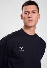 Čierne tričko s dlhým rukávom vyrobené z bavlny s okrúhlym výstrihom. Obsahuje malé biele vyšívané logo na ľavej časti hrude.