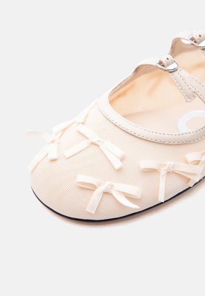 Μπεζ μπαλαρίνα με πολλά μικρά διακοσμητικά φιόγγους στη μύτη και ρυθμιζόμενο λουράκι με πόρπη στον αστράγαλο.