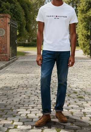 Mann trägt ein weißes Tommy Hilfiger T-Shirt, blaue Jeans und braune Wildlederschuhe und steht auf einem Kopfsteinpflasterweg in der Nähe eines Backsteintors.