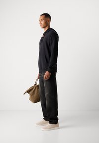 Schwarzer Hoodie mit einer Fronttasche, kombiniert mit dunklen Jeans und hellen Sneakers. Hält eine braune Tasche mit oberen Griffen.