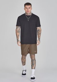 T-shirt noir à manches courtes avec une coupe décontractée, associé à un short de sport brun. Baskets noires et blanches, tatouages visibles sur les bras.