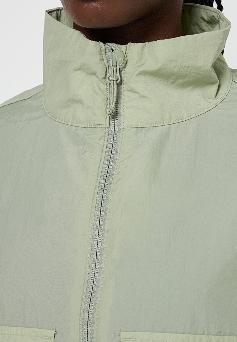 Veste zippée verte clair avec un col haut, en tissu texturé et deux poches avant. Présente une fermeture éclair centrale en nylon et des éléments de design minimalistes.
