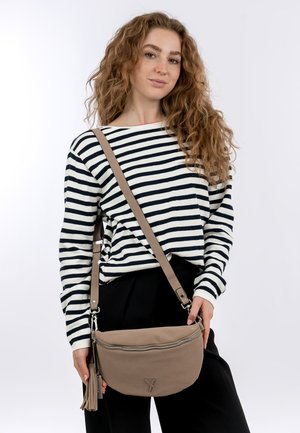 Ung kvinde med krøllet hår iført en sort-og-hvidstribet sweater og sorte bukser, der holder en beige crossbody-taske med frynse detaljer.