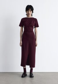 Burgunderfarbenes Kleid mit kurzen Ärmeln, tailliertem Oberteil und einem Seitenschlitz. Akzentuiert mit dunklen Slippern und passenden Socken. Glatte Stofftextur.