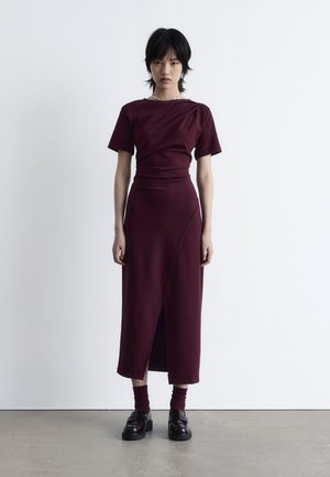 Burgundy dress with short sleeves, fitted bodice, and a side slit. Accented with dark loafers and matching socks. Smooth fabric texture.