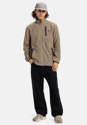 Veste en polaire marron clair avec fermeture éclair, mettant en avant des accents noirs et des poches, associée à un pantalon noir et des baskets grises. Style décontracté.