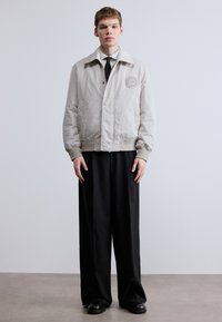 Ανοιχτό γκρι μπουφάν bomber με λογότυπο, διαθέτει φερμουάρ κλεισίματος και ριμπ μανσέτες, συνδυασμένο με φαρδιά μαύρη παντελόνια και μαύρα παπούτσια.