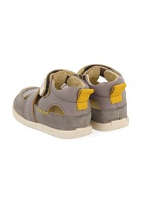 Sandalias grises y beige con una correa de velcro, puntera redondeada, acento amarillo en el talón y detalles de tela texturizada en los laterales.