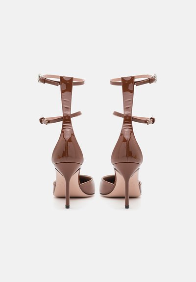 Zapatos de tacón alto de charol marrón con correas en los tobillos y soporte trasero, que presentan un diseño elegante y puntas afiladas.