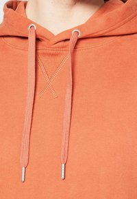 Orange hoodie med en mjuk textur, som har en framficka i känguruform och dragsnören med silverfärgade spetsar. Synlig sömnadsdetalj vid halsen.