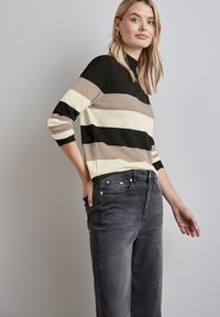 Schwarz-beiger und cremefarbener gestreifter Pullover mit langen Ärmeln und hohem Kragen, kombiniert mit dunkelgrauen Jeans mit vorderen Taschen.