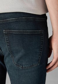 Vaqueros de mezclilla oscura con un corte clásico, que presentan un bolsillo trasero con costuras en contraste. La textura parece suave con un ligero desvanecimiento.