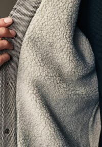 Grå og hvid jakke indvendig med blød fleece tekstur, med tæt vævet yderstof og synligt hardware til lukning.