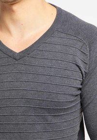 Homme portant un pull gris côtelé à col en V, montrant la partie supérieure de la poitrine et les épaules sur un fond blanc.