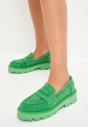 Pieds portant des mocassins en daim vert vif avec des semelles épaisses assorties de couleur verte sur un fond blanc.