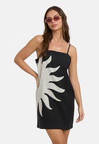 Vrouw in een zwart mouwloos jurkje met een groot wit zonmotief, met een roze getinte zonnebril en gelaagde gouden halskettingen, poseert met haar hand op haar heup.