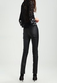 Kvinna i svart spets långärmad topp och åtsittande svarta läderbyxor, sedd bakifrån, stående mot en enkel ljus bakgrund.