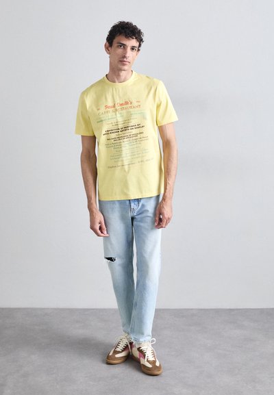 Yellow cotton t-shirt with printed text, light blue distressed jeans, and multi-colored sneakers with a suede and fabric combination.