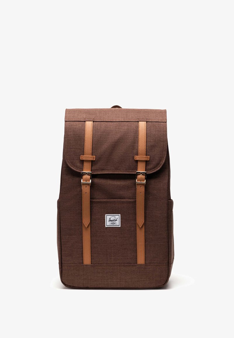 Ruskea kangasreppu, jossa on nahkaremmit ja etutasku, suorakulmainen muoto, läppäkiinnitys ja merkkilogo edessä.