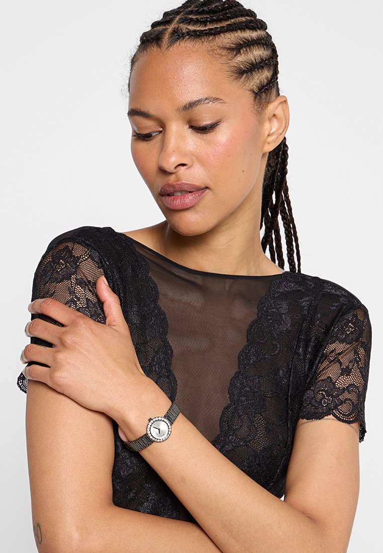 Frau mit geflochtenem Haar, die ein schwarzes Spitzenoberteil trägt, nach unten schaut, mit einer silbernen Armbanduhr am linken Handgelenk und den Arm über die Brust verschränkt.