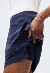 Πλάγια όψη ενός ατόμου που σηκώνει το κάτω μέρος μιας σκούρας μπλε φούστας-σορτς για να αποκαλύψει ενσωματωμένα σορτς από κάτω.