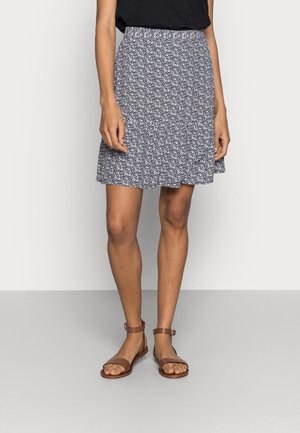 Frau trägt ein schwarzes Oberteil mit einem hoch taillierten blau-weißen Blumenrock und braunen Sandalen mit Knöchelriemen und steht auf einem grauen Boden.