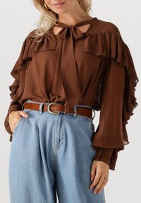 Vrouw in een bruin bloes met ruches en een strik om de hals, insteek in lichtblauwe high-waisted jeans met een brown riem, hand in de zak.