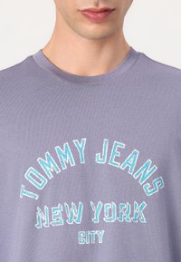 Violetinis medvilnės marškinėliai su siuvinėtu tekstu "TOMMY JEANS NEW YORK CITY" turkio ir baltos spalvos, apvaliu iškirpimu ir trumpomis rankovėmis.