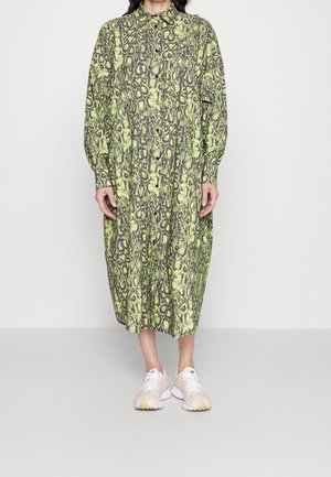 Kobieta w długiej sukience midi na guziki z długim rękawem, z zielono-czarnym wzorem wężowym, w białych trampkach, stojąca na jednolitym tle.