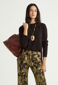 Maglione marrone lavorato a maglia, pantaloni a fantasia floreale in verde e beige, e una borsa bordeaux con texture intrecciata. Accessori comprendono una grande collana pendente.