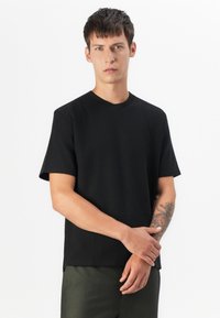 Camiseta negra de manga corta con un cuello redondo, hecha de algodón suave, que presenta un corte relajado y un diseño minimalista, combinada con pantalones verde oscuro.