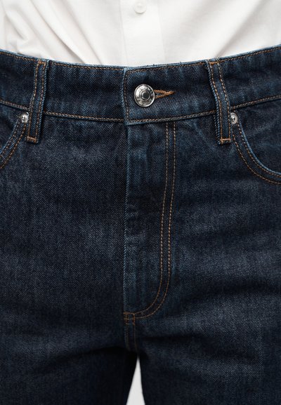 Κοντινό πλάνο στη μέση σκουρομπλέ τζιν παντελονιού με πορτοκαλί ραφές, μεταλλικό κουμπί και θηλιές ζώνης, φορεμένο με λευκό πουκάμισο που έχει μαζευτεί μέσα.