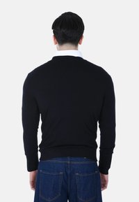 Uomo con capelli neri corti che indossa un maglione nero sopra una camicia bianca con colletto e jeans blu, rivolto di spalle contro uno sfondo semplice.