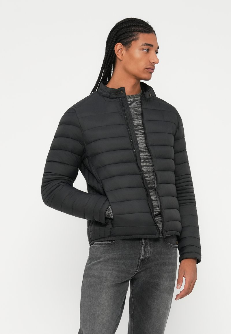 Doudoune noire avec col montant, design matelassé horizontal, fermeture éclair à l'avant et poches latérales. Portée par-dessus un pull gris.