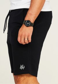 Black cotton shorts with drawstring waist, featuring a white logo on the hem. A black watch is worn on the wrist.