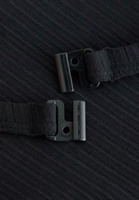 Musta kangas, jossa on kaksi mustaa solkea, kummankin pinta sileä ja kiiltävä, ja nauhat tekstuurisia, hillitysti raidallista taustaa vasten.