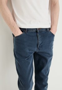 Jeans de denim azul escuro com um corte relaxado, apresentando um fecho de botão, bolsos frontais e detalhes de costura discretos na cintura.