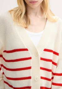 Pletená cardigan v krémovej farbe s červenými horizontálnymi pruhmi, s výstrihom do V, gombíkmi a textúrovanou väzbou. Mierne voľný strih.