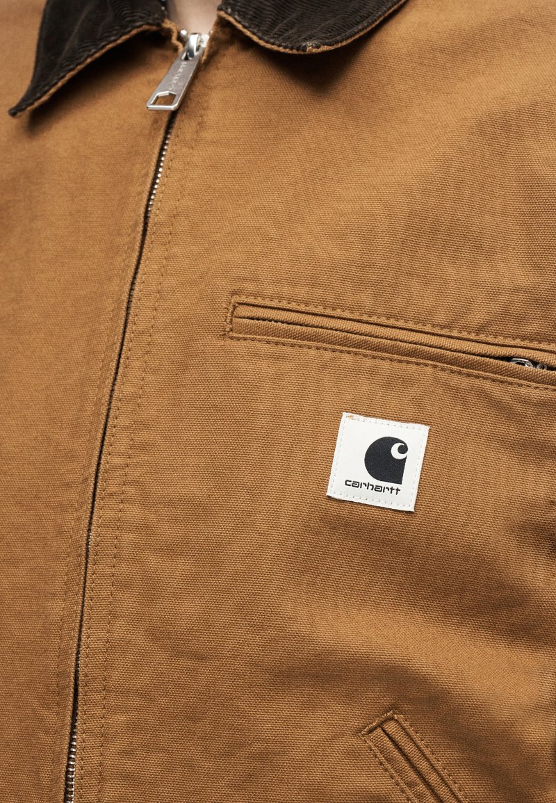 Brown zip-up jacket with dark collar, horizontal zip pocket, and white Carhartt logo patch on chest.