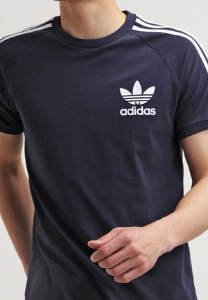 Osoba ubrana w granatowy T-shirt Adidas z białymi trzema paskami na rękawach i logo koniczyny Adidas na klatce piersiowej, ręka lekko uniesiona.