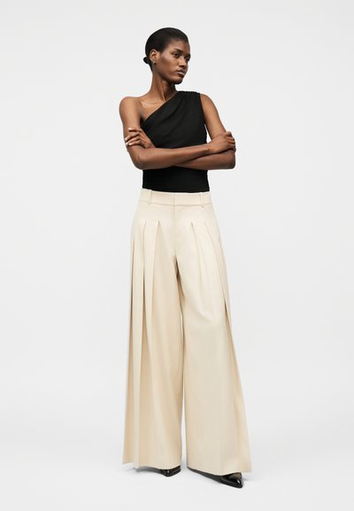 Μαύρη γυναίκα που στέκεται με σταυρωμένα τα χέρια, φοράει μαύρο τοπ με έναν ώμο και μπεζ πλατες παντελόνες με πιέτες, συνοδευόμενες από μαύρα μυτερά παπούτσια.