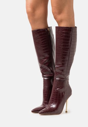 Stivali alti in pelle di coccodrillo borgogna con punta affusolata e tacchi a stiletto dorati indossati su gambe su uno sfondo bianco.